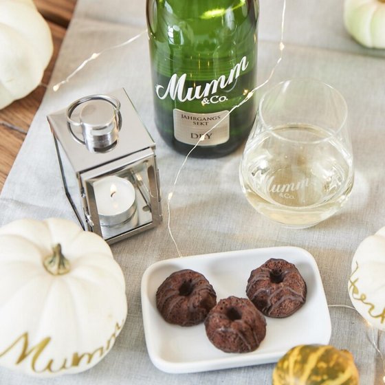 Ein eleganter Tisch mit einer Flasche Sekt, einem Glas, kleinen Schokoladenkuchen und dekorativen Kürbissen in sanften Farben.