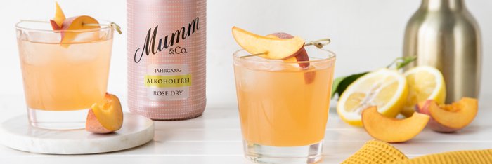 Auf dem Tisch liegen frische Zitronen und Pfirsiche, während zwei Gläser mit einem spritzigen Getränk im Vordergrund stehen.