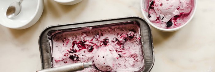 Eine Eisschale mit lila Beeren-Eis steht neben einer offenen Eisbox, die mit einem Löffel bearbeitet wird.