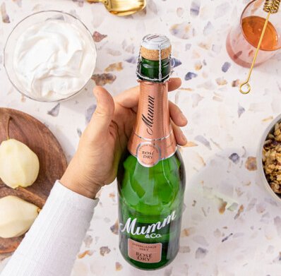 Die Flasche Mumm Rosé Dry steht im Mittelpunkt, während eine Hand sie greift, umgeben von frischen Birnen und anderen Leckereien.