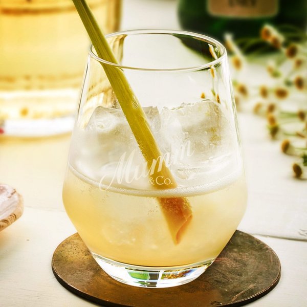 Das Glas zeigt ein köstliches, helles Getränk mit Eiswürfeln und einem dekorativen Zitronengras-Stängel, umgeben von Blumen.