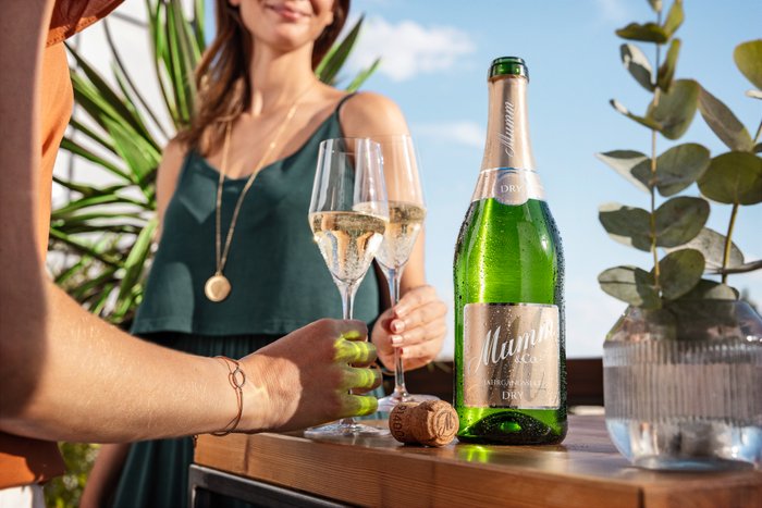 Eine grüne Flasche steht neben zwei mit Sekt gefüllten Gläsern und einer Frau im Hintergrund