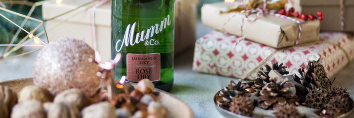 Eine Flasche Mumm Sekt steht auf einem Tisch, umgeben von Geschenken und festlichen Dekorationen wie Tannenzapfen und Kugeln.