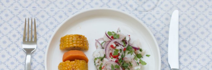 Ein Teller mit zwei Stücken Mais, einer Karotte und einem frischen Salat aus Zwiebeln und Koriander auf einem gemusterten Tisch.