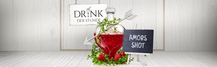 Ein herzförmiger Glasbehälter mit rotem Getränk, umgeben von frischen Erdbeeren und grünen Pflanzen, steht auf einem hellen Tisch.