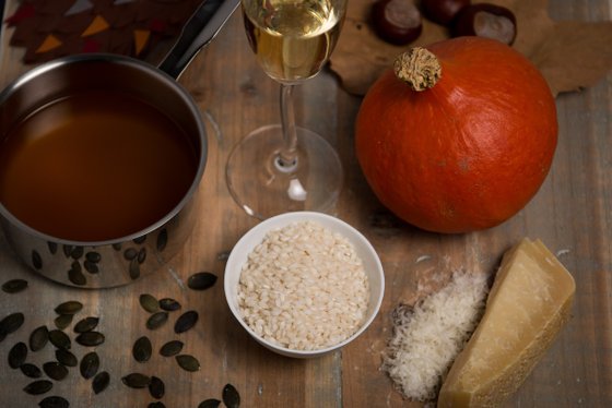 Auf einem rustikalen Tisch sind ein Topf mit Brühe, ein Kürbis, Reis und geriebener Käse kunstvoll angeordnet.