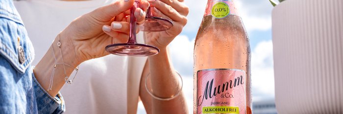 Eine Frau mit Sonnenbrille stößt mit einem Glas mit einer andere Person mit blauem Hemd an, daneben steht eine rosane Flasche Sekt und eine Pflanze