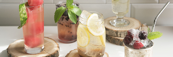 Auf einem hellen Tisch sind verschiedene Cocktails arrangiert, darunter ein fruchtiger Drink mit Beeren und ein klarer, sprudelnder Cocktail.