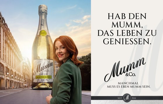Vor einer malerischen Stadtansicht präsentiert eine Frau mit einem charmanten Lächeln eine große Flasche alkoholfreien Sekt von Mumm.