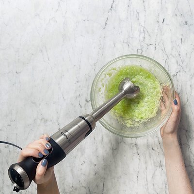 Eine Person hält einen Stabmixer über eine Schüssel mit grünem Püree auf einer marmorierten Oberfläche.