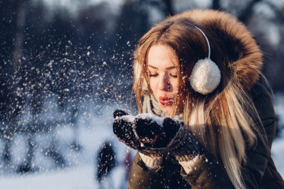 Eine junge Frau mit Kopfhörern bläst fröhlich Schnee in die Luft, während sie in einer verschneiten Landschaft steht.