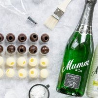 Zwei Flaschen Mumm Sekt liegen neben einer Anordnung von Schokoladenkugeln auf einem weißen Untergrund.