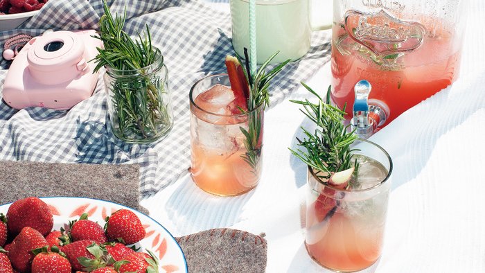 Drei Gläser mit erfrischenden Getränken, garniert mit Rosmarin, stehen auf einem karierten Tuch neben frischen Erdbeeren.