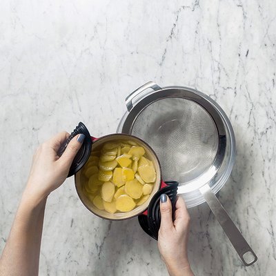 Hände einer Person, die einen Topf mit eingelegten Gurken über ein Sieb auf einem hellen Marmortisch halten.
