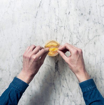 Zwei Hände greifen nach einer Zitronenscheibe, die in einem Glas mit orangefarbenem Getränk schwimmt, auf einer marmorierten Oberfläche.