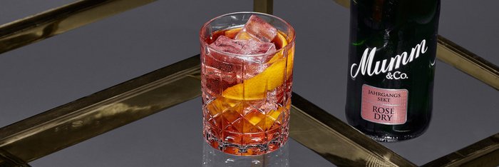 Auf einem stilvollen Tisch steht ein Glas mit einem erfrischenden rotfarbigen Getränk, das Eis und eine Orangenscheibe enthält, neben einer Sektflasche.