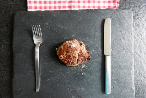 Ein saftiges Stück Steak liegt auf einem schwarzen Schieferbrett, umgeben von einem Messer und einer Gabel.