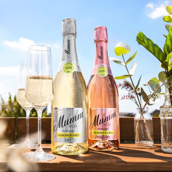 Auf einem Holztisch stehen zwei Sektflaschen, einmal rosa einmal gelblich, daneben zwei gefüllte Sektgläser und im Hintergrund ein sonniger Himmel