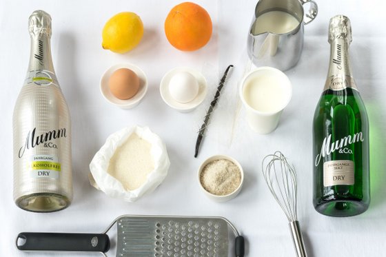 Auf einem weißen Tisch liegen Zutaten für ein Rezept: alkoholfreier Sekt, Eier, Zitrusfrüchte und verschiedene Milchprodukte.