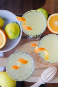 Auf einem Holzbrett stehen drei Gläser mit einem gelben Cocktail, dekoriert mit Orangenschalen und umgeben von Zitronen und Orangen.