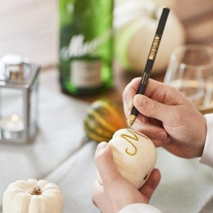 Eine Person bemalt einen kleinen weißen Kürbis mit einem goldenen Buchstaben, umgeben von herbstlichen Dekorationen.