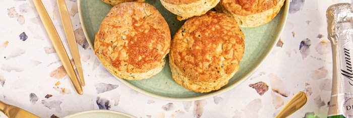 Die Szene zeigt köstliche Scones auf einem grünen Teller, begleitet von goldenen Bestecken und einer Flasche Sekt.
