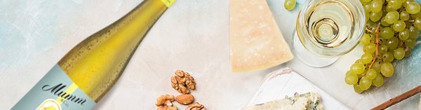 Eine Flasche Mumm-Sekt steht neben einem Glas, umgeben von Trauben, Nüssen und verschiedenen Käsesorten auf einem hellen Tisch.