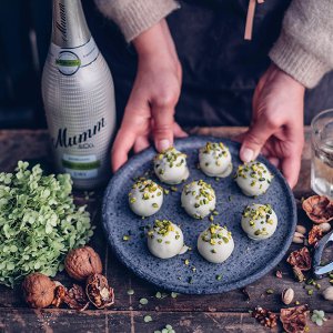 Auf einem rustikalen Tisch stehen Pralinen auf einem blauen Teller, während eine Flasche Sekt und Nüsse im Hintergrund sichtbar sind.