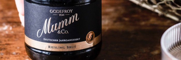 Eine Flasche Godefroy von Mumm Riesling Brut steht auf einem rustikalen Holztisch, umgeben von sanften Lichtreflexen.