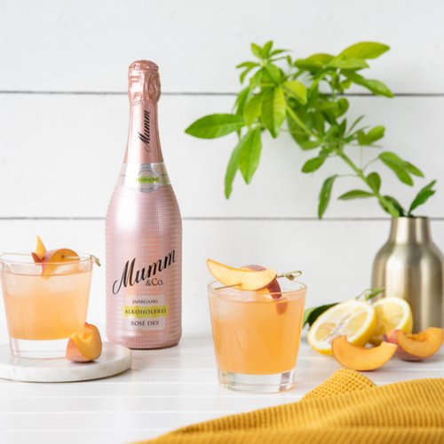 Zwei Gläser mit einem erfrischenden, roséfarbenen Getränk, garniert mit Pfirsichscheiben, stehen neben einer Flasche Mumm.