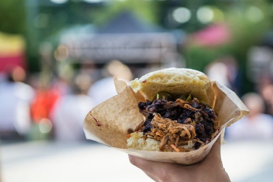 Eine Hand hält ein Wrap mit zartem, zerrissenem Fleisch und frischen Zutaten, während eine lebhafte Veranstaltung im Hintergrund stattfindet.