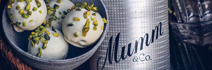 Eine Schüssel mit weißen Pralinen, garniert mit gehackten Pistazien, steht neben einer Flasche Mumm & Co.
