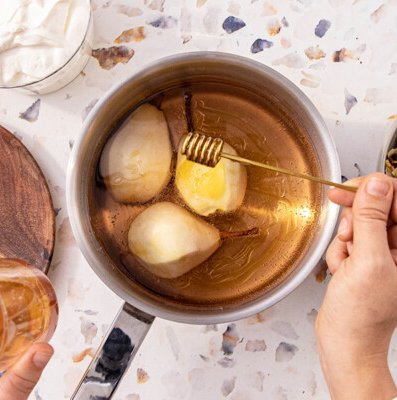In einem Topf werden drei Birnen in einer goldenen Flüssigkeit gekocht, während ein Löffel Honig darüber geträufelt wird.