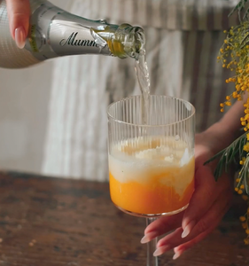 Ein Glas mit orangefarbenem Getränk wird mit sprudelndem Sekt gefüllt, während eine Hand das Glas hält.