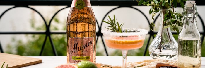 Auf einem Tisch am Wasser liegen eine Flasche Mumm, ein Cocktail mit Grapefruit und Limette sowie frische Kräuter und Sonnenbrille.