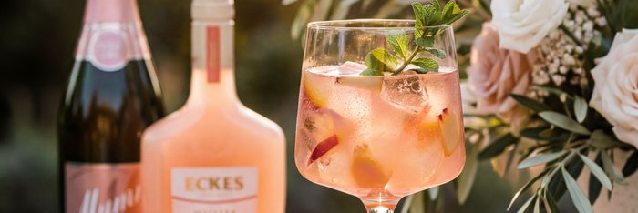 Ein White Peach Spritz Aperitif mit den Flaschen Eckes White Peach & Mumm & Co Rosé Dry im Hintergrund