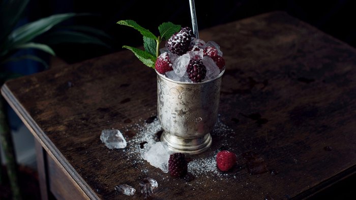 Ein silberner Becher gefüllt mit Eis, Himbeeren und Brombeeren, steht auf einem dunklen Holztisch, umgeben von Eisstücken.