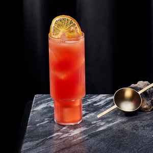 Ein erfrischender, roter Cocktail mit Eiswürfeln, präsentiert in einem stilvollen Glas neben einem goldenen Messlöffel.