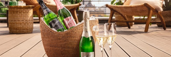 Auf einem Rillen-Boden steht eine Sektflasche und zwei Sektglser sowie dahinter ein Sektkühler, im hinteren Hintergrund ist eine Sommerterrasse zu antizipieren