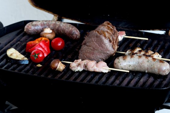 Auf dem Grill liegen verschiedene Fleischsorten, darunter Würste und Spieße, umgeben von buntem Gemüse wie Paprika und Tomaten.