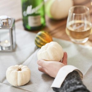 Eine Hand hält zwei kleine, weiße Kürbisse, während ein Glas Wein und eine Flasche im Hintergrund sichtbar sind.