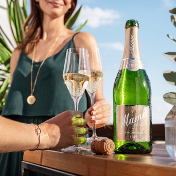 Eine grüne Flasche steht neben zwei mit Sekt gefüllten Gläsern und einer Frau im Hintergrund