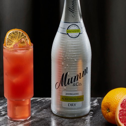Eine Flasche Mumm alkoholfrei steht neben einem bunten Cocktail und frischen Zitrusfrüchten auf einem marmorierten Tisch.