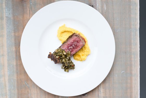 Ein saftiges Stück Rindfleisch liegt auf einem weißen Teller, garniert mit einer würzigen Kräutersauce und cremigem Polenta.