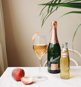Ein stilvolles Arrangement mit einem Glas Rosé-Sekt, einer Flasche Mumm und einem frischen Pfirsich auf einem weißen Tisch.