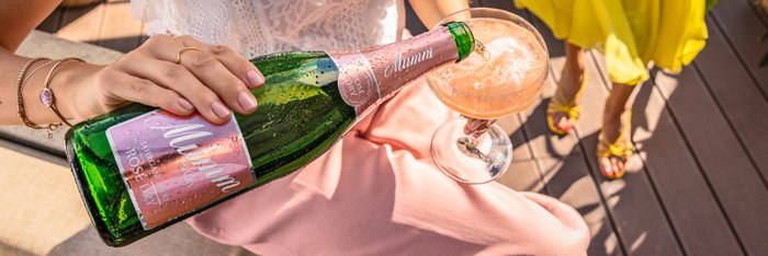 Eine Frau schenkt Sekt aus einer grünen Sektflasche mit rosa Etikett in ein Glas ein, das auf einem Tisch mit rosa Tuch steht.