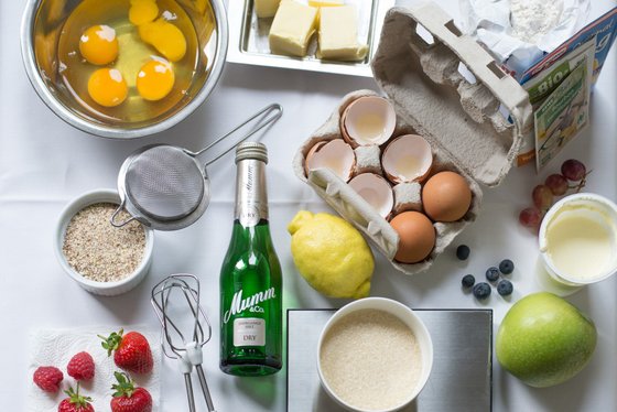 Eine Vielzahl von Zutaten liegt auf einem Tisch, darunter frische Eier, Obst und eine Flasche Sekt, bereit zum Kochen.