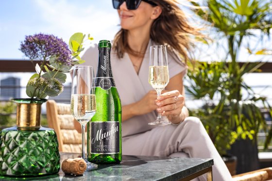 Im Hintergrund sitzt eine Frau mit Sonnenbrille und einem Glas in der Natur, im Vordergrund steht eine Flasche Mumm Sekt auf einem Tisch