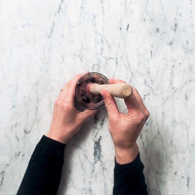 Das Bild zeigt Hände, die einen Holzstößel in ein Glas mit dunklen Beeren drücken, auf einem eleganten Marmortisch.