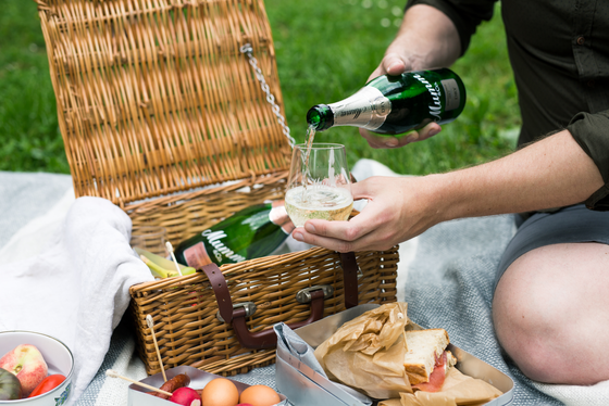 Ein Mann gießt Sekt in ein Glas, während er auf einer Decke im Freien sitzt, umgeben von einem Picknickkorb und frischen Lebensmitteln.